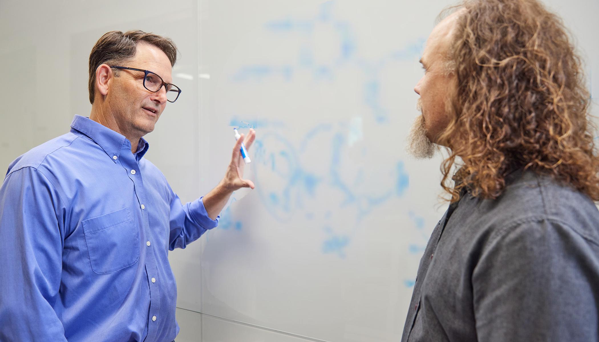 博彩平台网址大全 scientists Brett Crawford and Dan Wendt talking by a whiteboard with a scientific formula